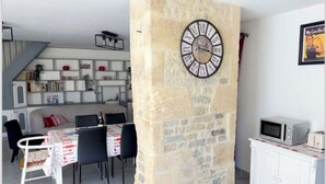 Una sala moderna con un pilar de piedra, un comedor con mesa y sillas blancas, una zona de cocina con microondas, y una estantería.