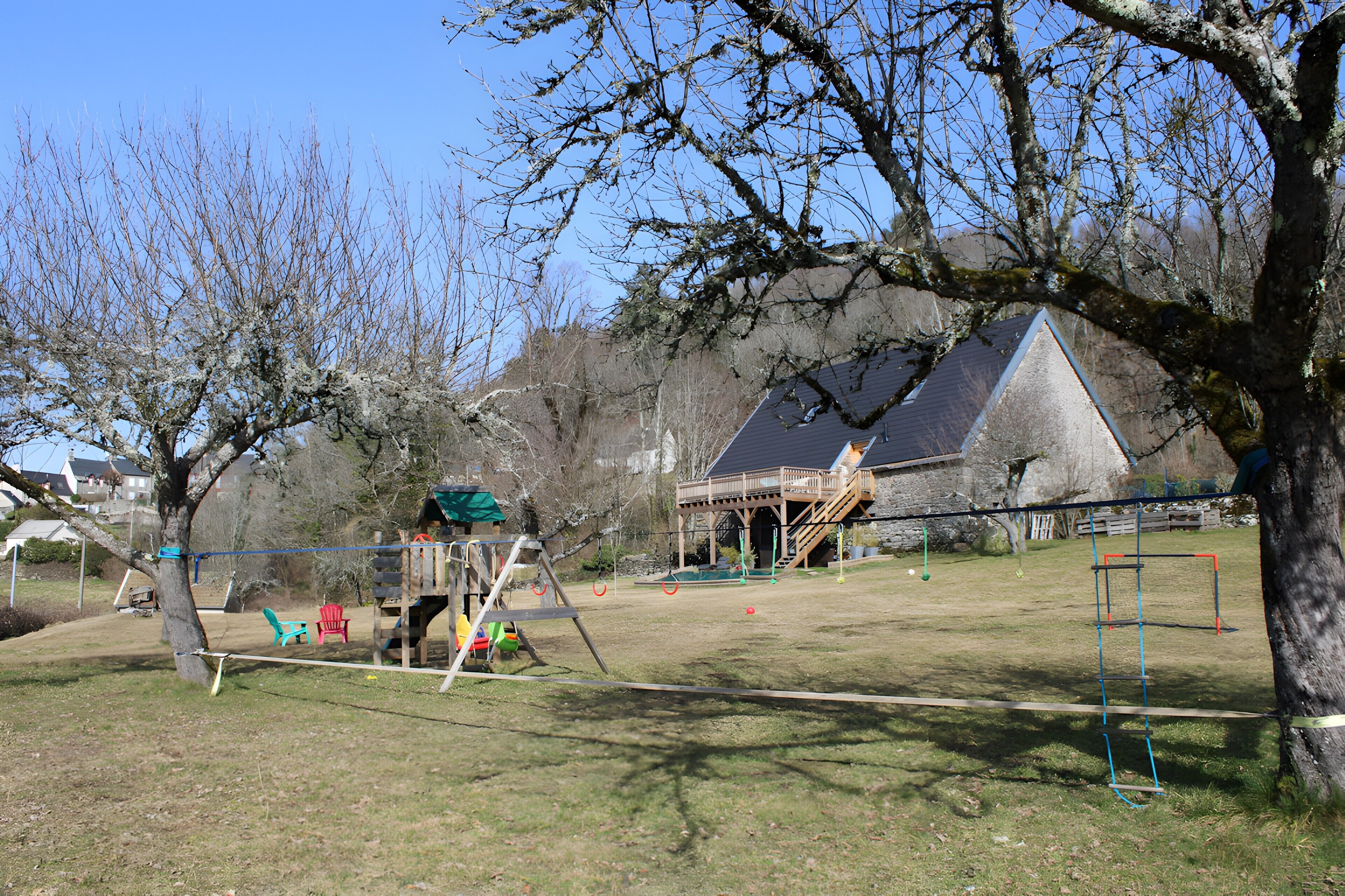 Un terrain de jeux avec des balançoires et des toboggans, une terrasse en bois et, au fond, une maison avec un toit très pentu.