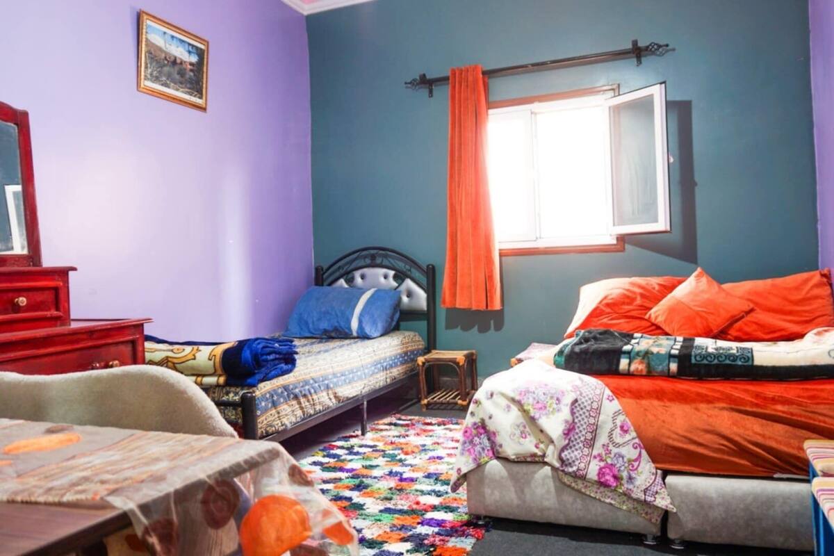 Quarto com duas camas, uma cômoda, uma janela com cortinas e um tapete colorido.