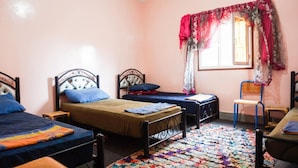 A bedroom with two single beds, a window with a red curtain, a chair, and a colorful rug. - Auberge de la jeunesse d ouarzazate  (Tarmigt)