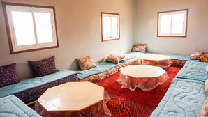 A room with multiple seating areas, including sofas and a central table, with a red patterned rug and light blue bedding. - Auberge de la jeunesse d ouarzazate  (Tarmigt)