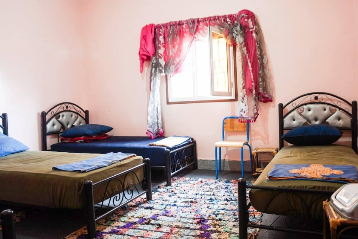 Quarto com duas camas de solteiro, uma mesinha, uma cadeira e janela com cortina.