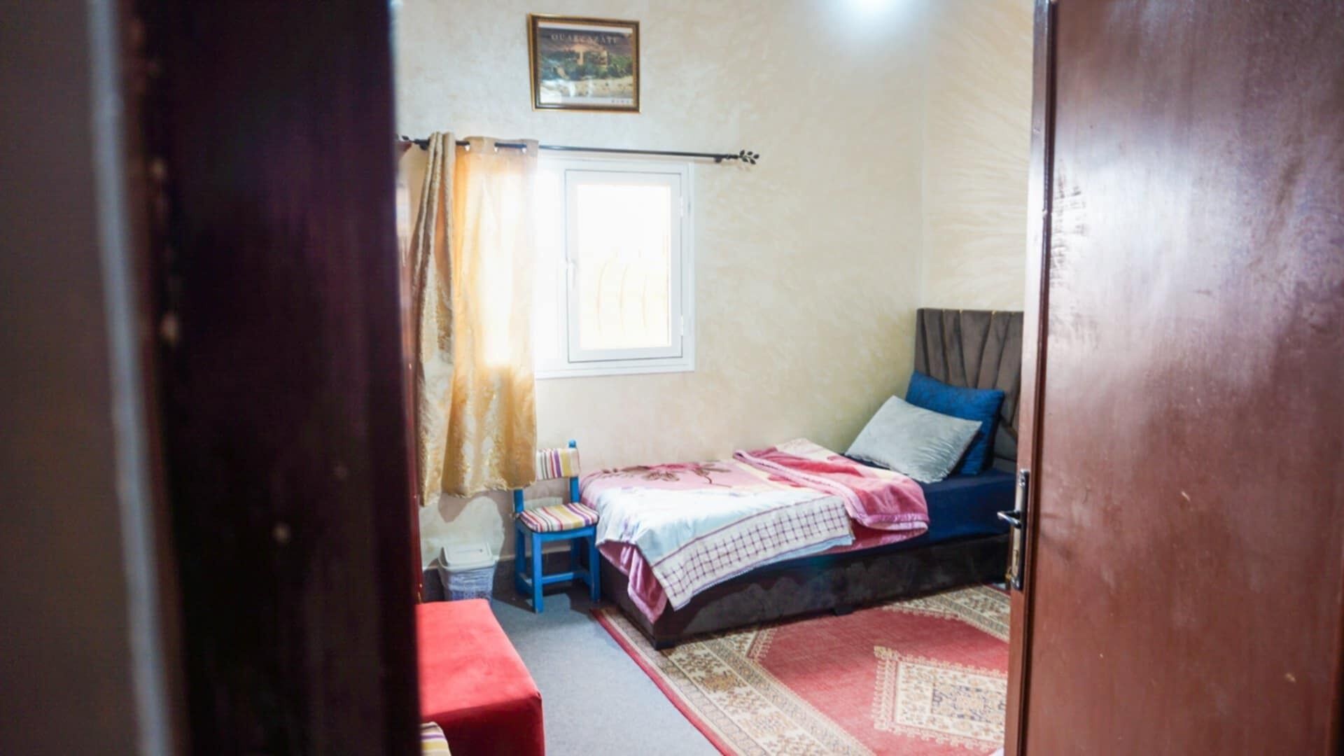 Une chambre à coucher avec un lit, une fenêtre avec des rideaux, un petit tabouret et un tapis rouge.