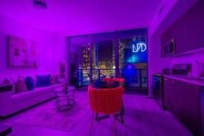 A modern living room with a sofa, a red chair, and a dining table. There is a large window with a city view and a neon sign. - Brickell Area| Free Parking| Balcony| Rooftop Pool (Miami)