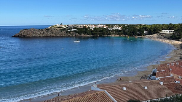 A coastal view with a beach, a rocky outcrop, and a small sailboat. - Apartment 'Dreams Of Menorca' with Sea View, Shared Pool, and Wi-Fi (Arenal des Castell)
