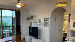 A modern living room with a television, dining table, chairs, a ceiling fan, and a washing machine. - Apartment 'Dreams Of Menorca' with Sea View, Shared Pool, and Wi-Fi (Arenal des Castell)