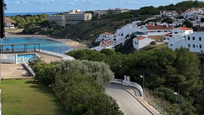 A coastal town with a pool, a curved road, and buildings with red roofs. - Apartment 'Dreams Of Menorca' with Sea View, Shared Pool, and Wi-Fi (Arenal des Castell)