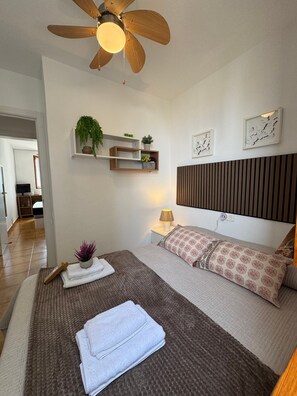 Un dormitorio con cama, mesitas de noche, ventilador de techo, estantes en la pared con plantas y una lámpara de noche.
