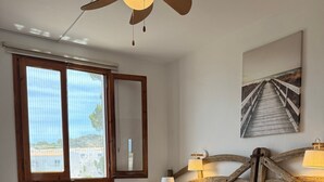 A neatly made bed with pillows, a bedside table with a lamp, a ceiling fan, a window with blinds, and a wall-mounted picture. - Apartment 'Dreams Of Menorca' with Sea View, Shared Pool, and Wi-Fi (Arenal des Castell)
