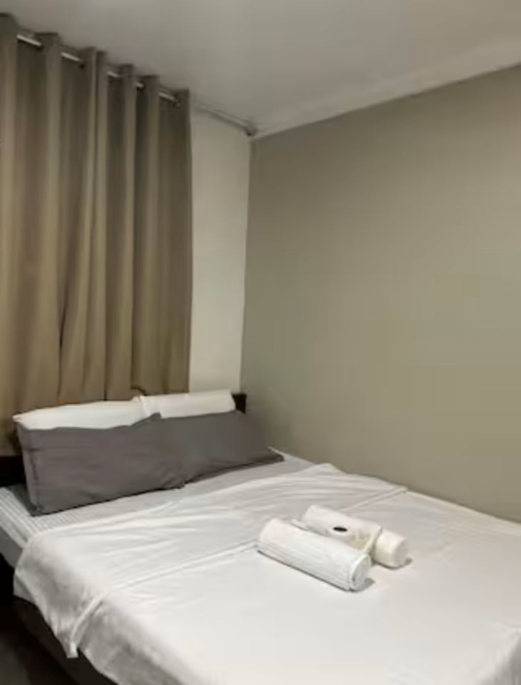 A neatly made bed with white linens and a grey pillow, a window with beige curtains, and a plain beige wall.