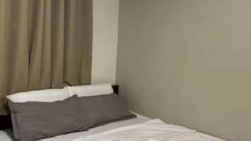 A neatly made bed with white linens and a grey pillow, a window with beige curtains, and a plain beige wall.