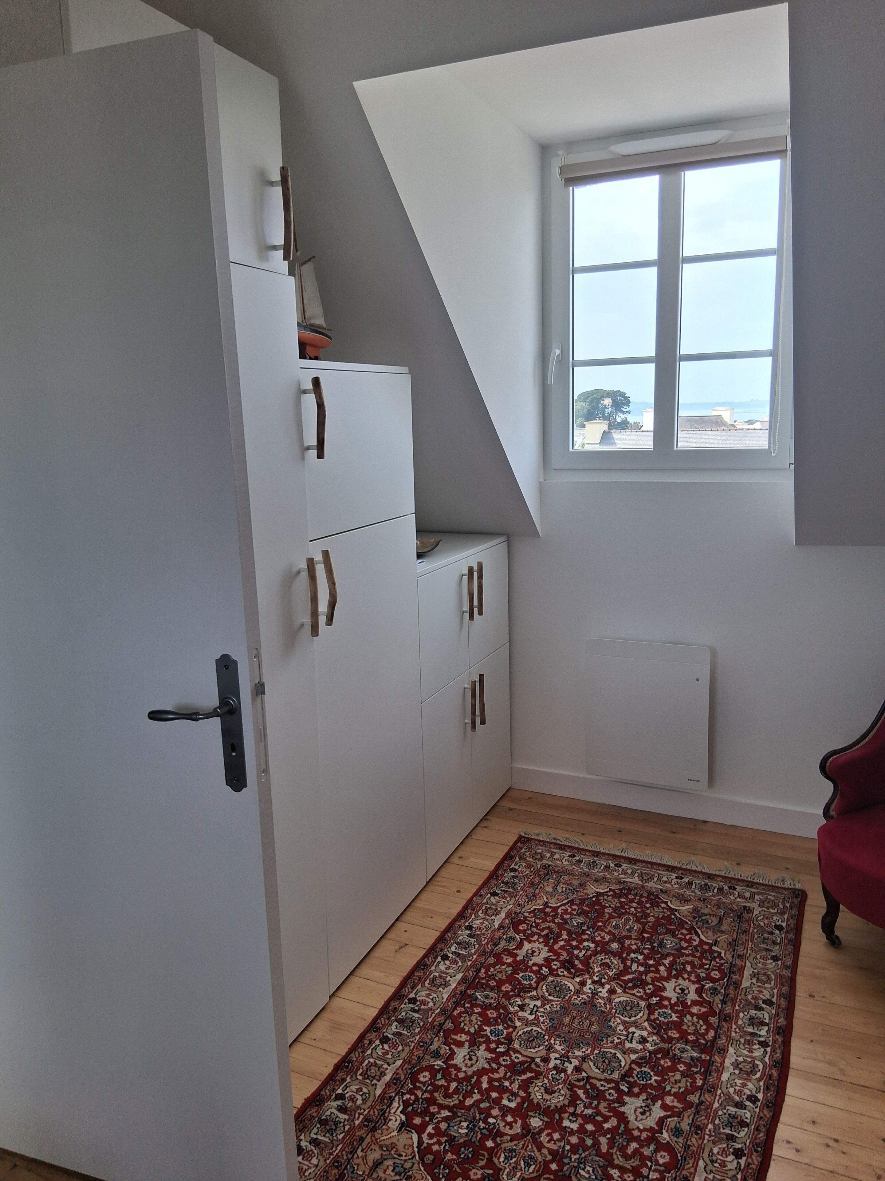 Une pièce avec une armoire blanche, un tapis à motifs, une fenêtre donnant sur un paysage agréable et un radiateur.