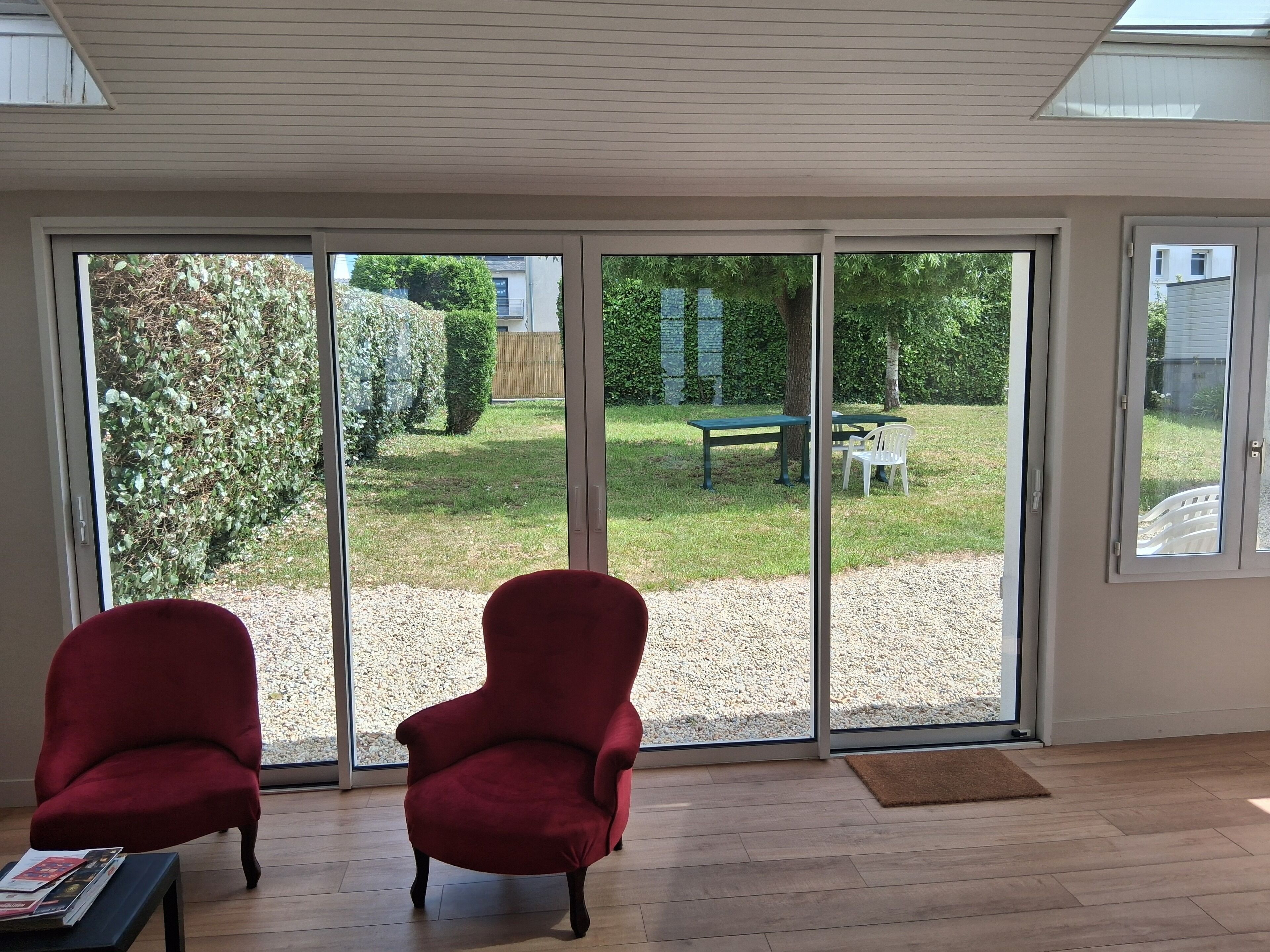 Ein Zimmer mit zwei roten Sesseln, Holzboden und Blick durch große Schiebeglastüren auf einen Garten mit Bank und Tisch.