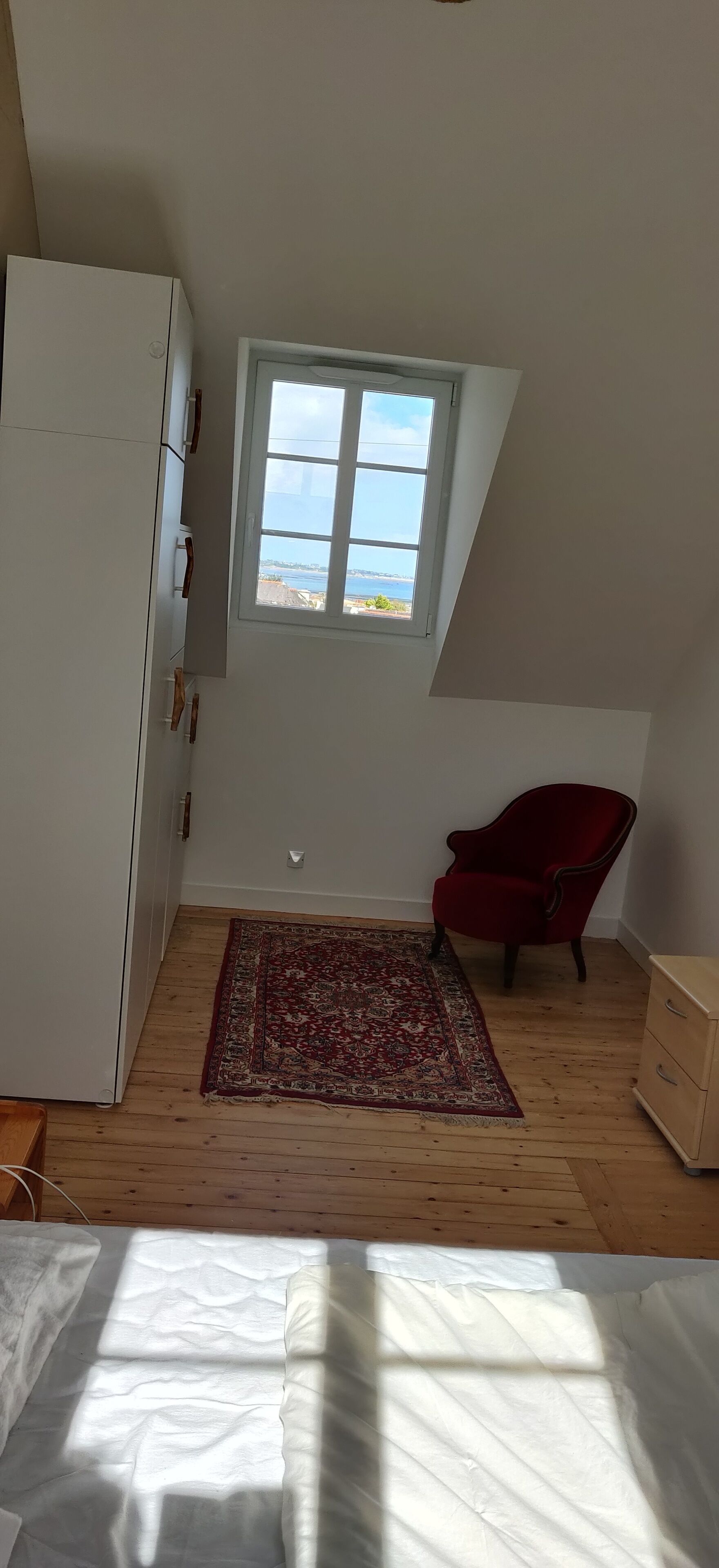 A room with a sloped ceiling, a window with a view, a red chair, a wooden floor, and a white cabinet.