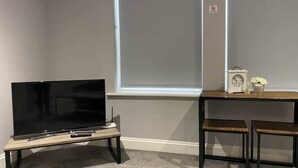 Una habitación con televisor sobre un soporte de madera, una mesita con un reloj y flores, y dos sillas.
