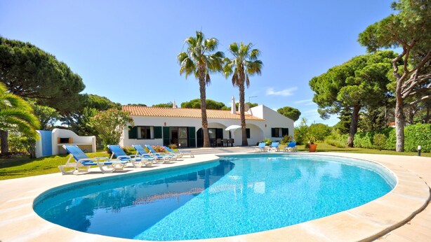Una piscina con sillas de descanso, una casa con techo de tejas y palmeras.