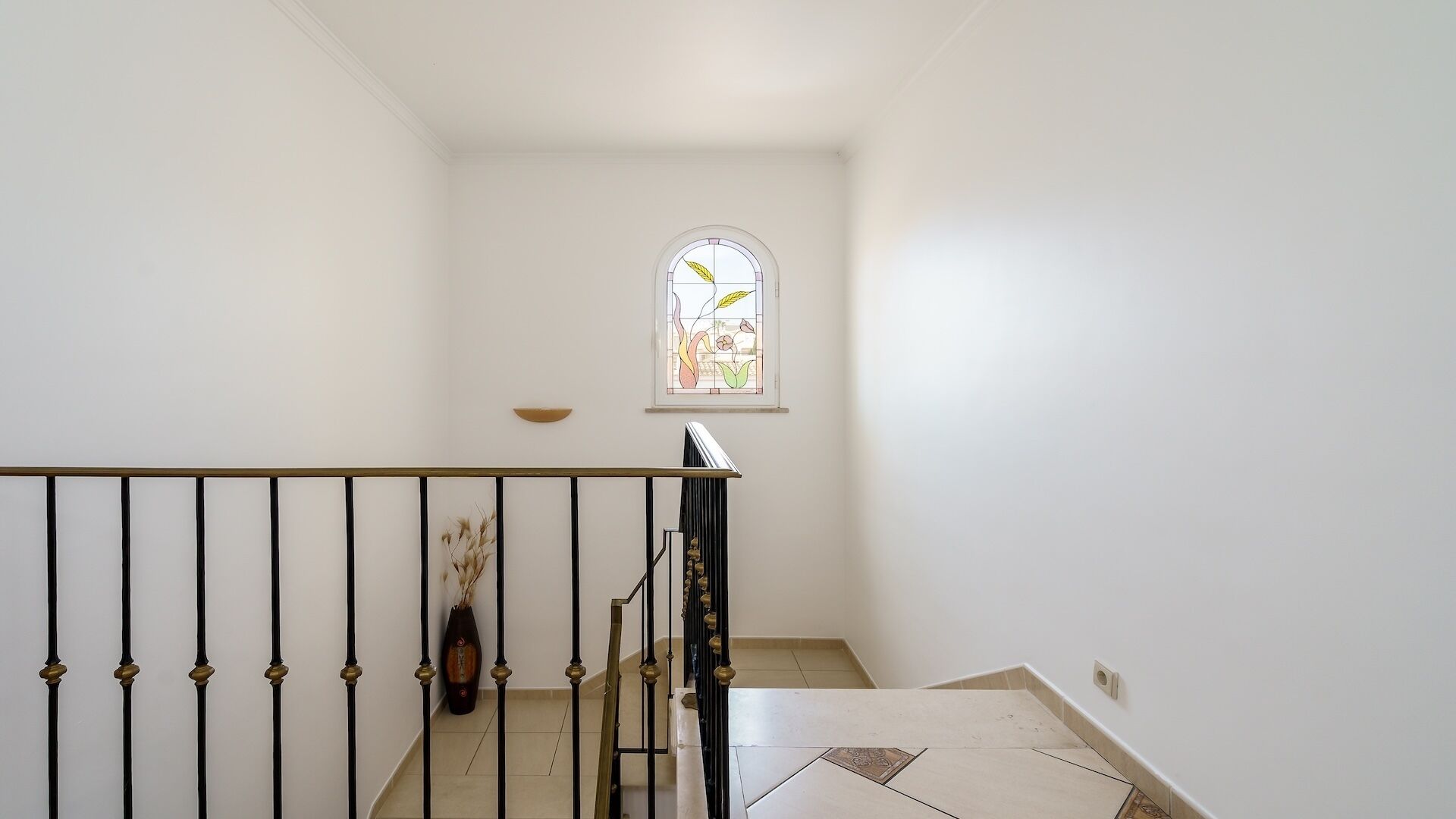 Un escalier avec une balustrade noire, un petit vase décoratif accroché au mur et une fenêtre ornée de vitraux.
