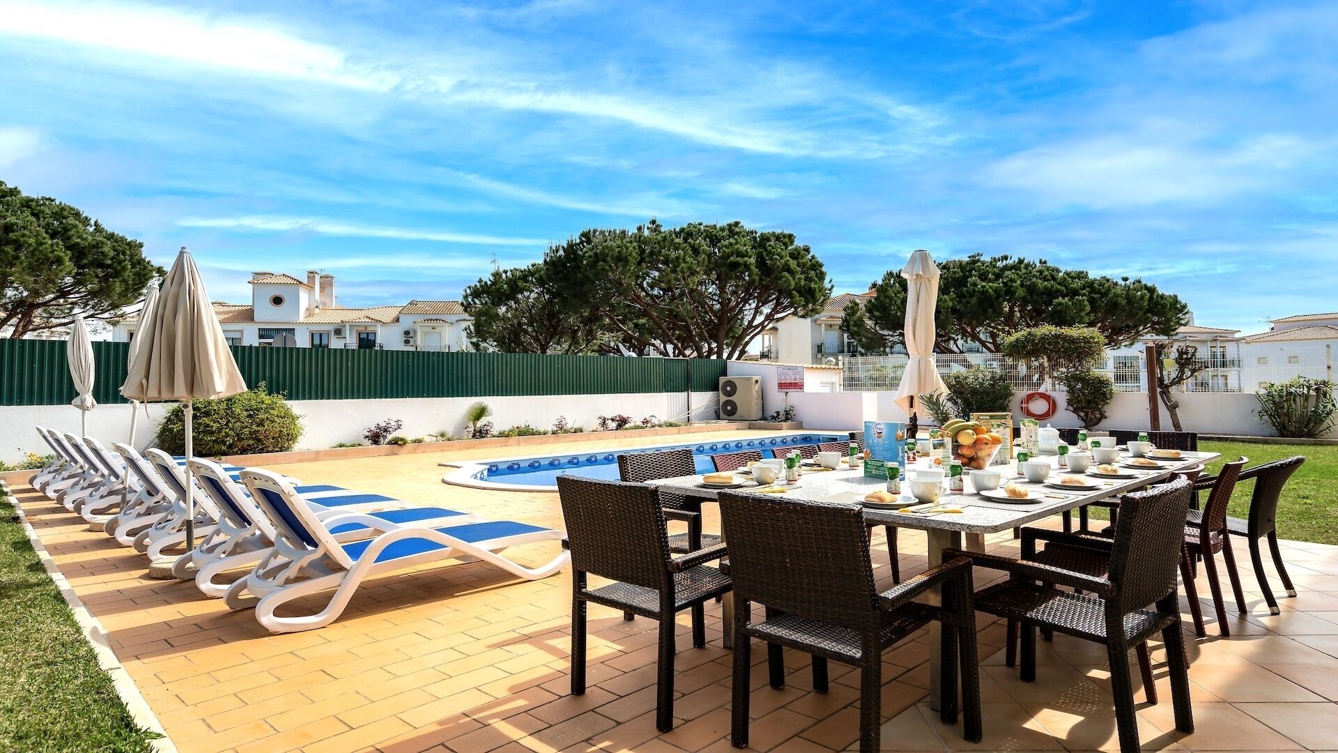 Un área de comedor junto a la piscina con sillones de descanso y una mesa dispuesta para una comida, rodeada de una piscina y follaje.
