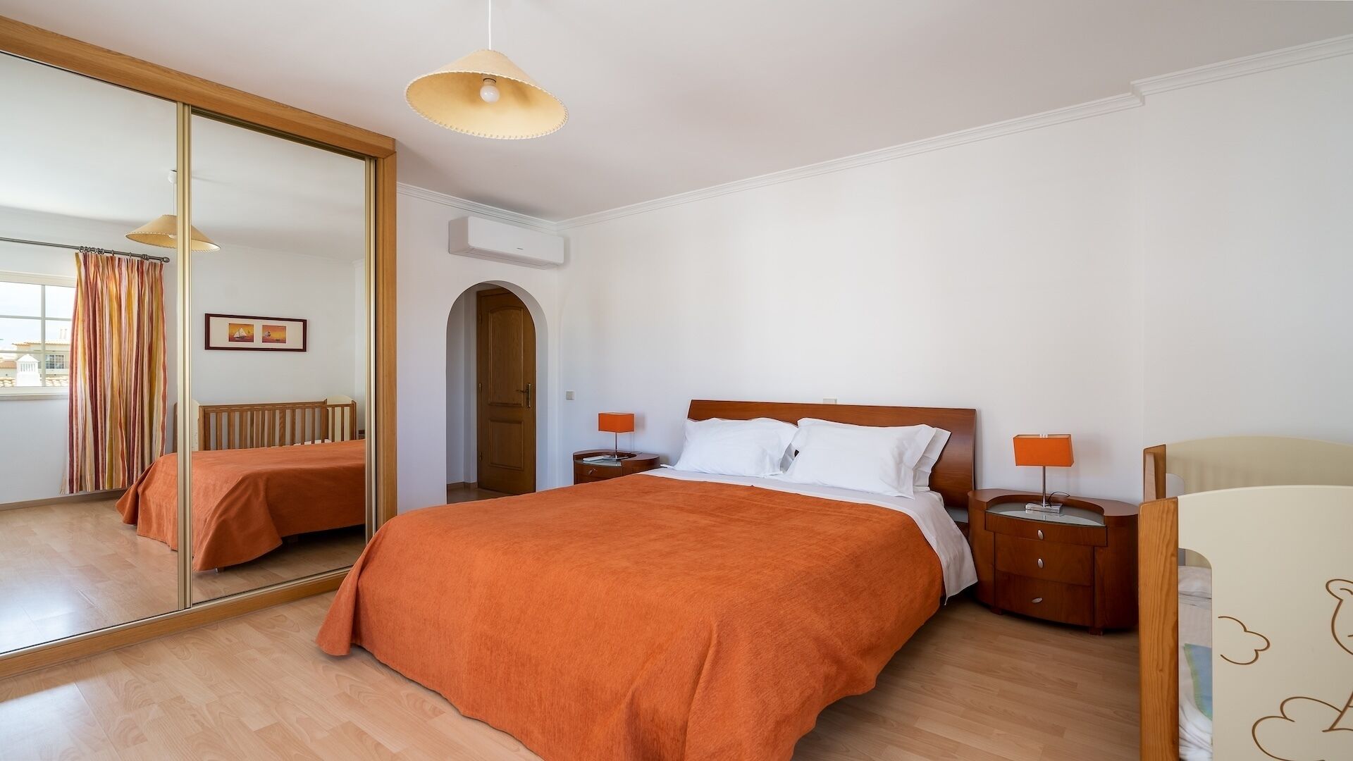 Une chambre d’hôtel avec un grand miroir, un lit en bois recouvert d’une couverture orange, deux tables de chevet avec des lampes et une armoire en bois.