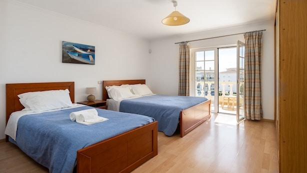 A hotel room with two wooden beds, a painting of boats, a lamp, and a view of the sea through a glass door. - Villa Brady (Olhos de Água)