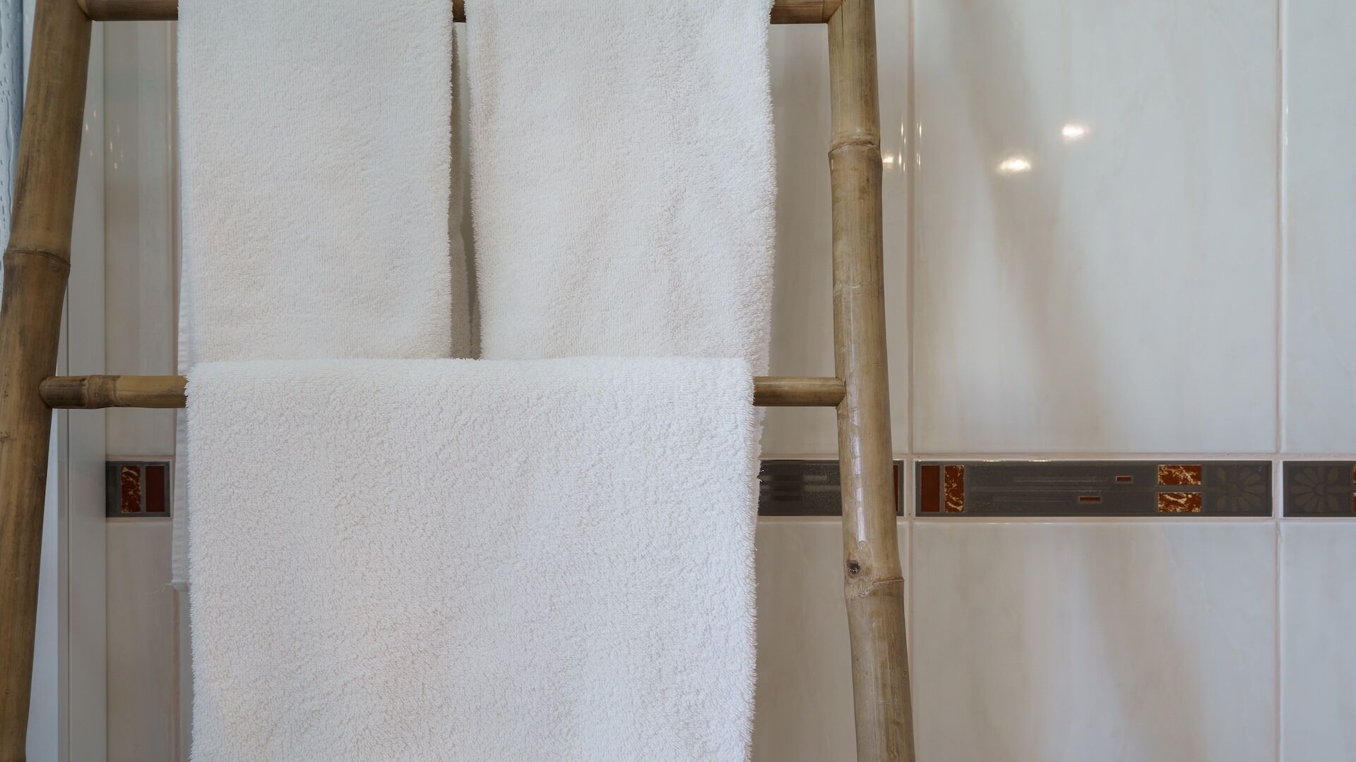 A white towel hanging on a bamboo ladder in a bathroom with white tiles and decorative borders.