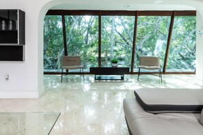 Una sala de estar moderna con una mesa de centro de vidrio, dos sillones, un sofá y un ventanal con vistas a árboles.