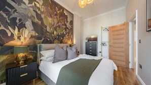 Ein Schlafzimmer mit einer großen floralen Wandmalerei, einem Bett mit vielen Kissen, einem Nachttisch mit Lampe und einem Kleiderschrank mit Spiegel.