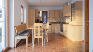 A modern kitchen with wooden cabinets, a dining table with chairs, a bench, and a built-in oven. - ღLiboria | Bright balcony apartment by the lake (Riegsee)