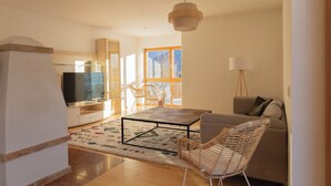A modern living room with a sofa, a coffee table, and a TV. There are wicker chairs and a lamp. - ღLiboria | Bright balcony apartment by the lake (Riegsee)
