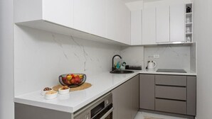 Une cuisine compacte avec des meubles blancs, un pare-feu en marbre et un petit plan de travail agrémenté d’un panier de fruits et de pots.