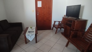 Een woonkamer met een donkergrijze bank, een klein houten tafeltje met een kantkleed, een televisie op een houten dressoir, en een deur met een berichtje erop.