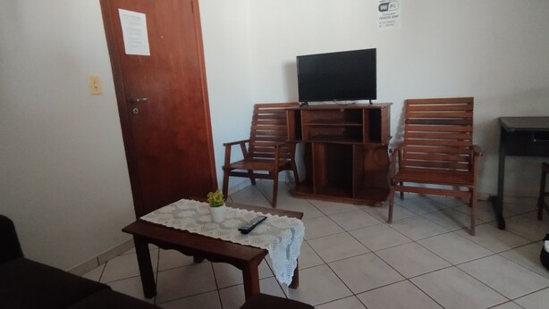 Een kleine kamer met een houten tv-standaard, twee houten stoelen, een klein tafeltje met een afstandsbediening, en een deur met een bordje.