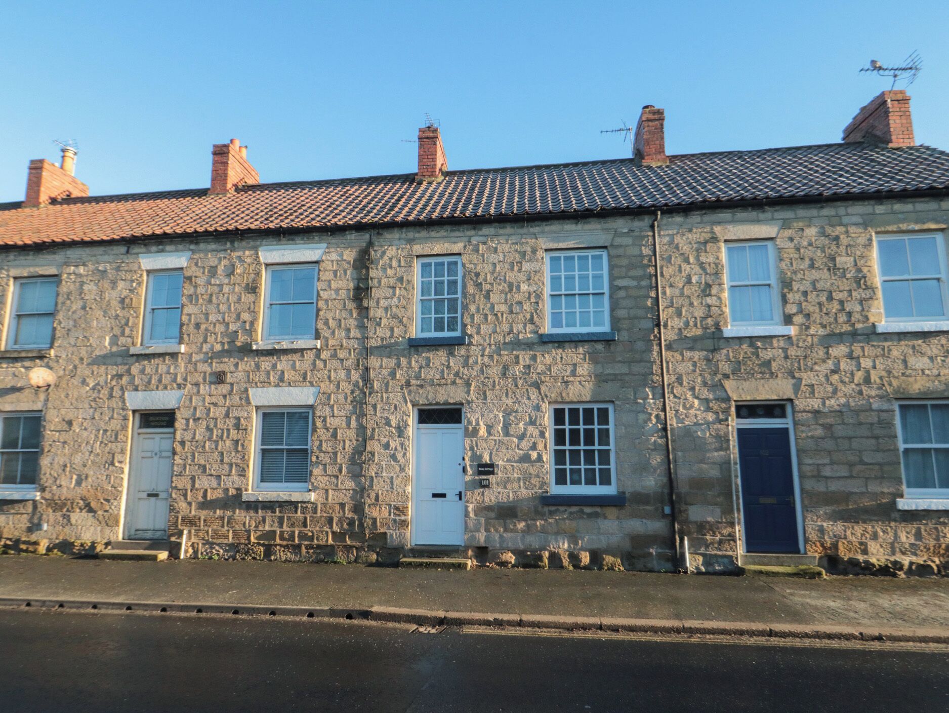 A row of stone buildings with multiple windows and doors, a paved street in front, and a clear blue sky.