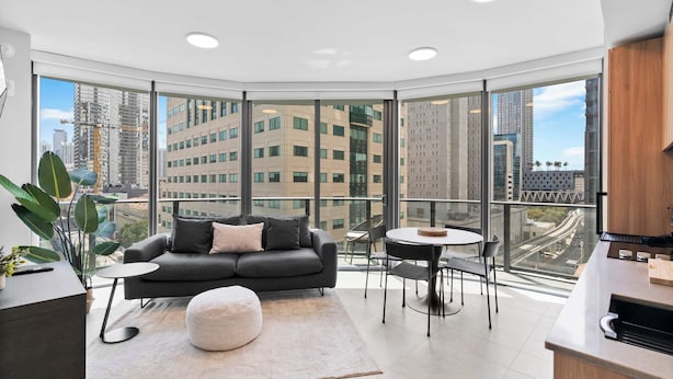 Un salon moderne avec un canapé, un pouf et une table ronde, de grandes fenêtres offrant une vue sur la ville, et un coin cuisine.