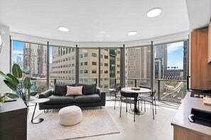 A modern living room with a sofa, ottoman, and round table, large windows with a city view, and a kitchen area. - Luxury 1BR | Free Parking | Brickell 5 Min (Miami)