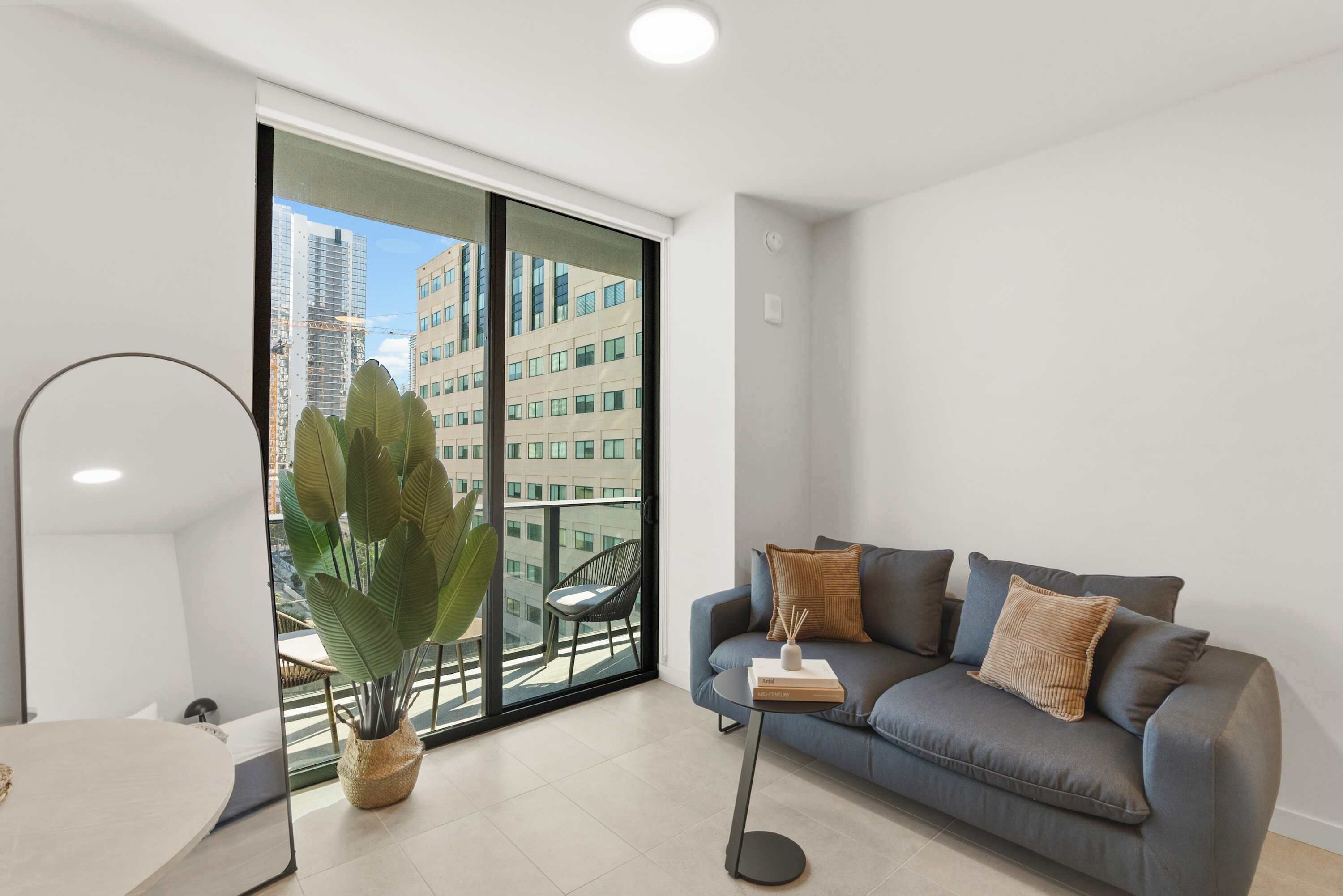 A modern living room with a sofa, a small table, a potted plant, and a view of buildings through a large window.