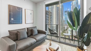A modern living room with a grey sofa, a round coffee table, and a large window offering a view of high-rise buildings. - Stylish 1BR+Den | Pool / CoWork | Free Parking (Miami)