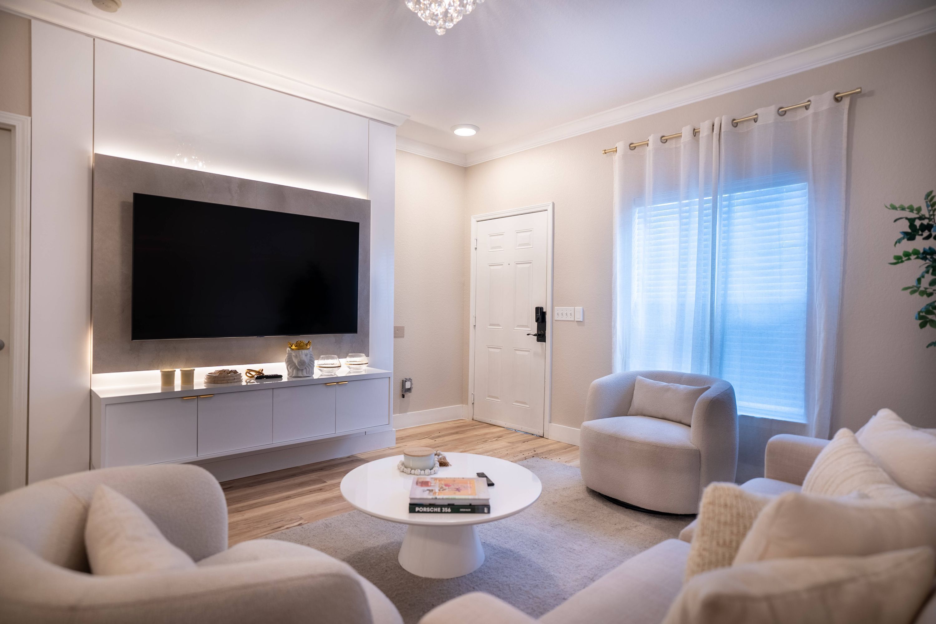 Una sala de estar moderna con una televisión de pantalla plana, sofás blancos, una mesa de centro y un amplio ventanal con cortinas.
