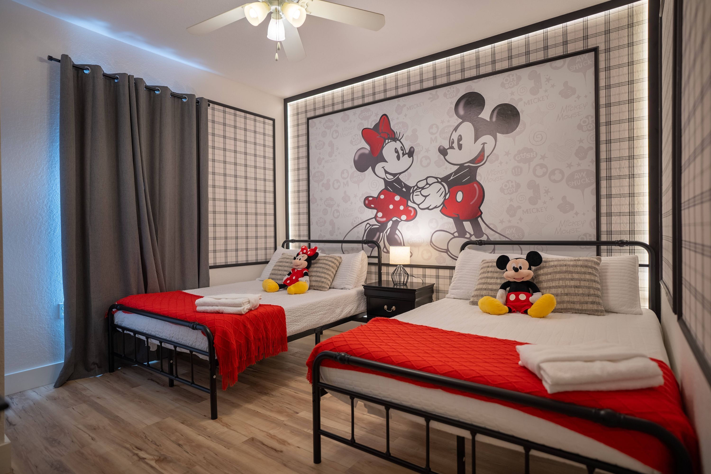 A hotel room with two beds, each with a Mickey Mouse plush toy, a red blanket, and a Mickey Mouse-themed wall mural.