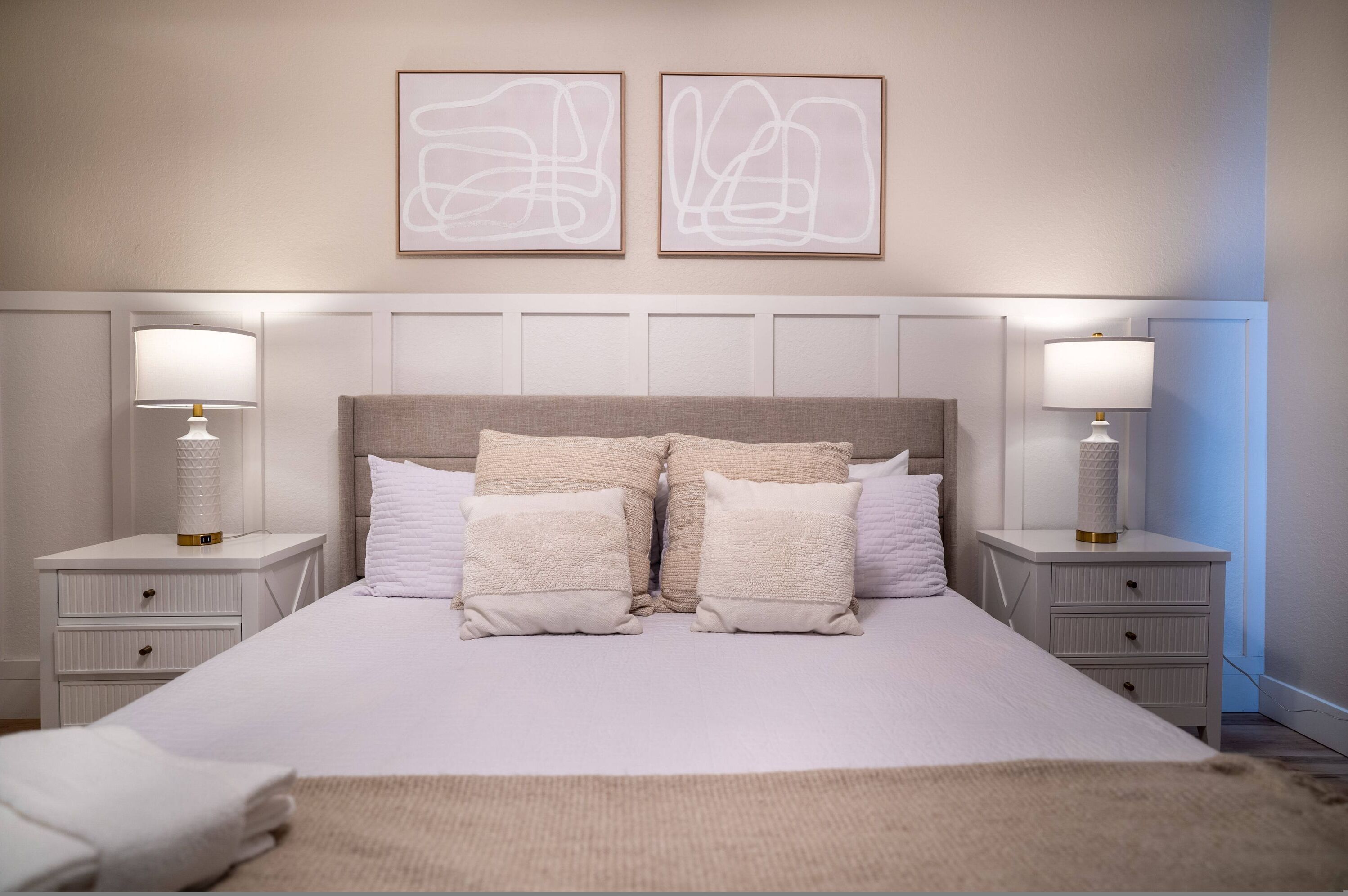 Un dormitorio ordenado con una cama grande, dos mesitas de noche y dos cuadros abstractos colgados en la pared.