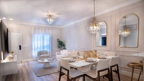 Uma sala de jantar com uma mesa branca posta para quatro pessoas, rodeada por cadeiras, com um candelabro acima.