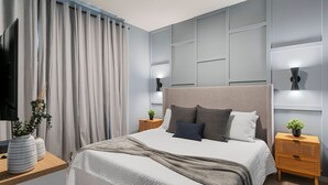 Un dormitorio moderno con una cama grande, cortinas grises, un televisor de pantalla plana, lámparas de noche y una planta pequeña en la mesita de noche.