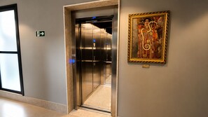 Un ascensor con pantalla digital, un letrero de salida de emergencia en la pared y una obra de arte enmarcada.