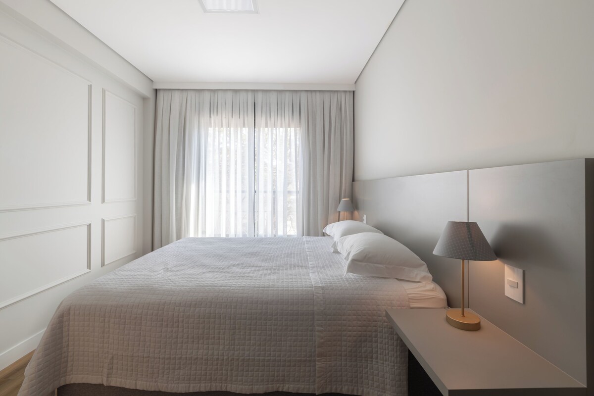 A neatly made bed with a white duvet, a bedside lamp, and a window with sheer curtains.