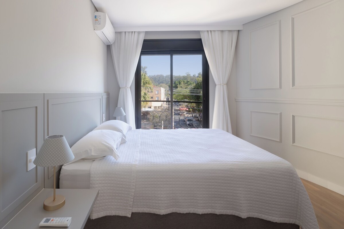 A bedroom with a bed, a bedside table with a lamp, a remote control, and a window with curtains.