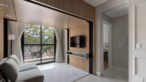 A modern living room with a sofa, a flat-screen TV mounted on a wooden wall, a large window with curtains, and a hallway leading to another room. - 1986 Apparts  (Águas de São Pedro)