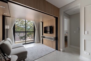 Ein modernes Wohnzimmer mit einer Couch, einem Flachbildfernseher an einer Holzwand, einem großen Fenster mit Vorhängen und einem Flur, der in einen anderen Raum führt.