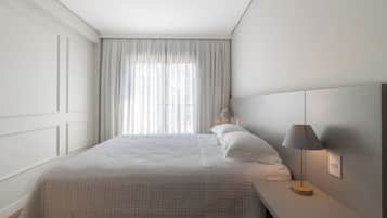 Una cama bien hecha con edredón blanco, una lámpara de noche y una ventana con cortinas translúcidas.