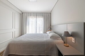 Un letto rifatto con un piumone bianco, una lampada da comodino e una finestra con tende leggere.