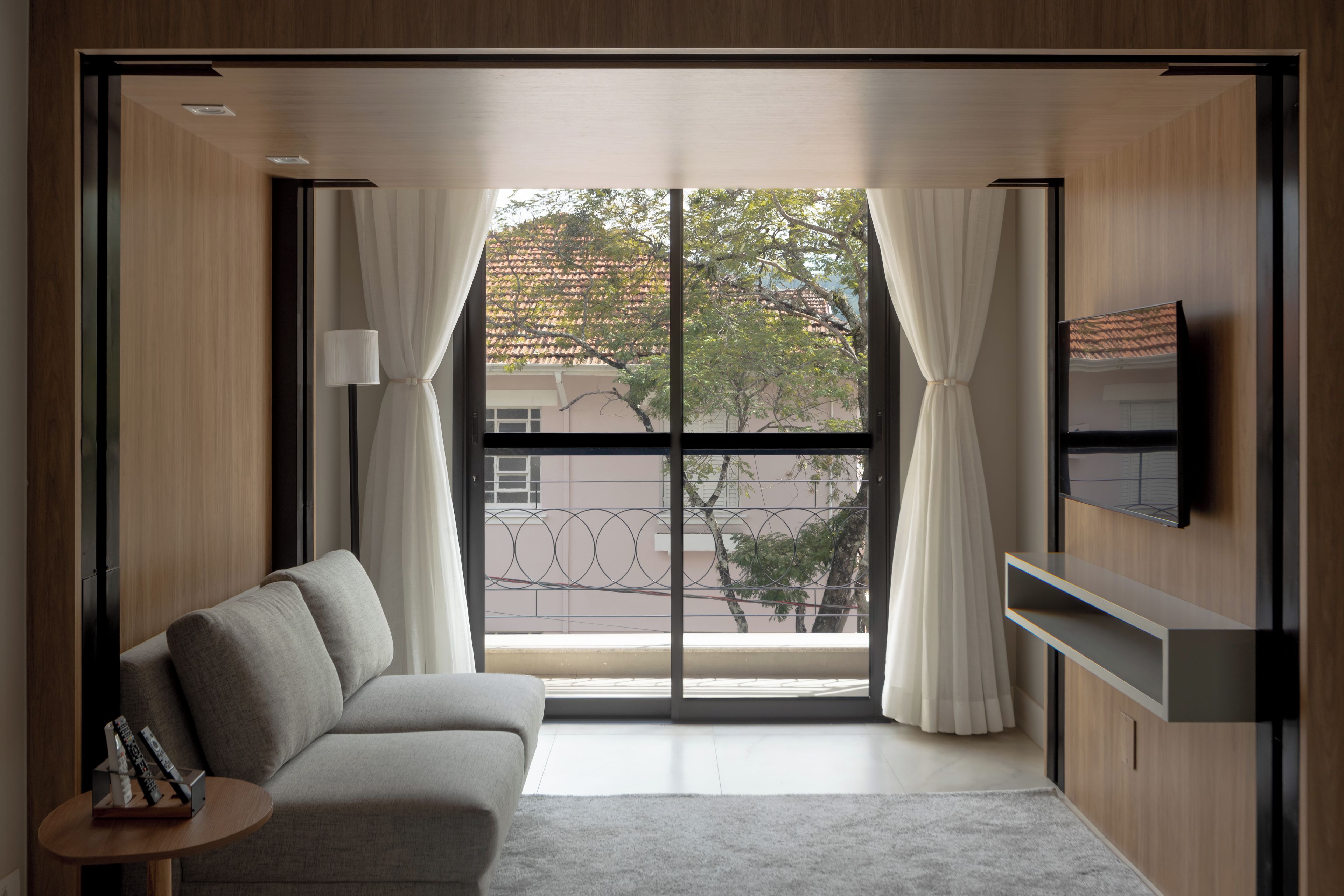 A modern living room with a sofa, a TV mounted on the wall, and a large window with curtains.